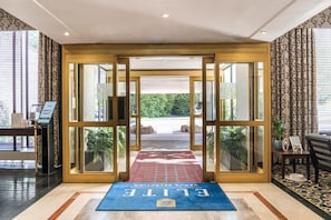 Entrada de un hotel con puerta de cristal, alfombra roja y felpudo azul con el nombre del hotel.
