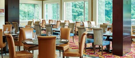 A spacious dining area with multiple tables and chairs, large windows with a view of trees, and a patterned carpet.
