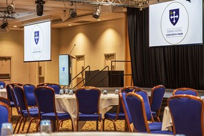 A conference room with rows of blue chairs, a stage with a podium, and a large screen displaying a logo. - Grand Hotel Gosforth Park (Newcastle-upon-Tyne)