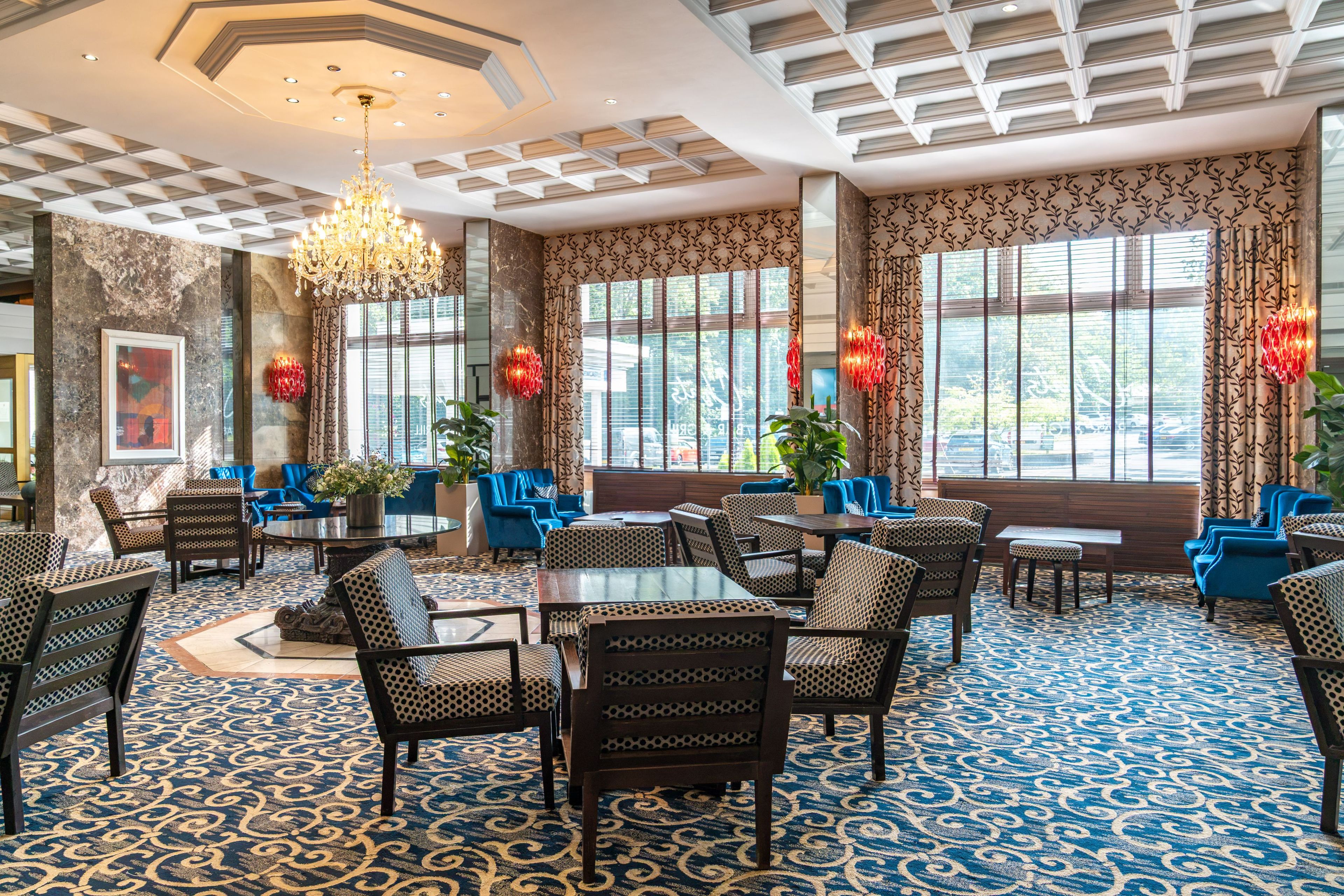 A hotel lobby with a patterned carpet, blue upholstered chairs, a chandelier, and large windows with curtains.