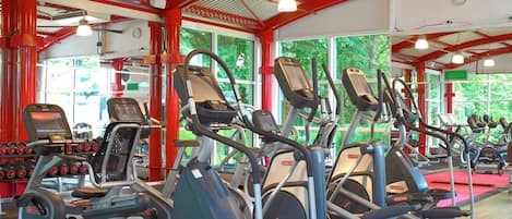 Une salle de sport équipée de plusieurs tapis de course et d’appareils elliptiques, avec des poutres rouges apparentes et de grandes fenêtres donnant sur un paysage verdoyant.