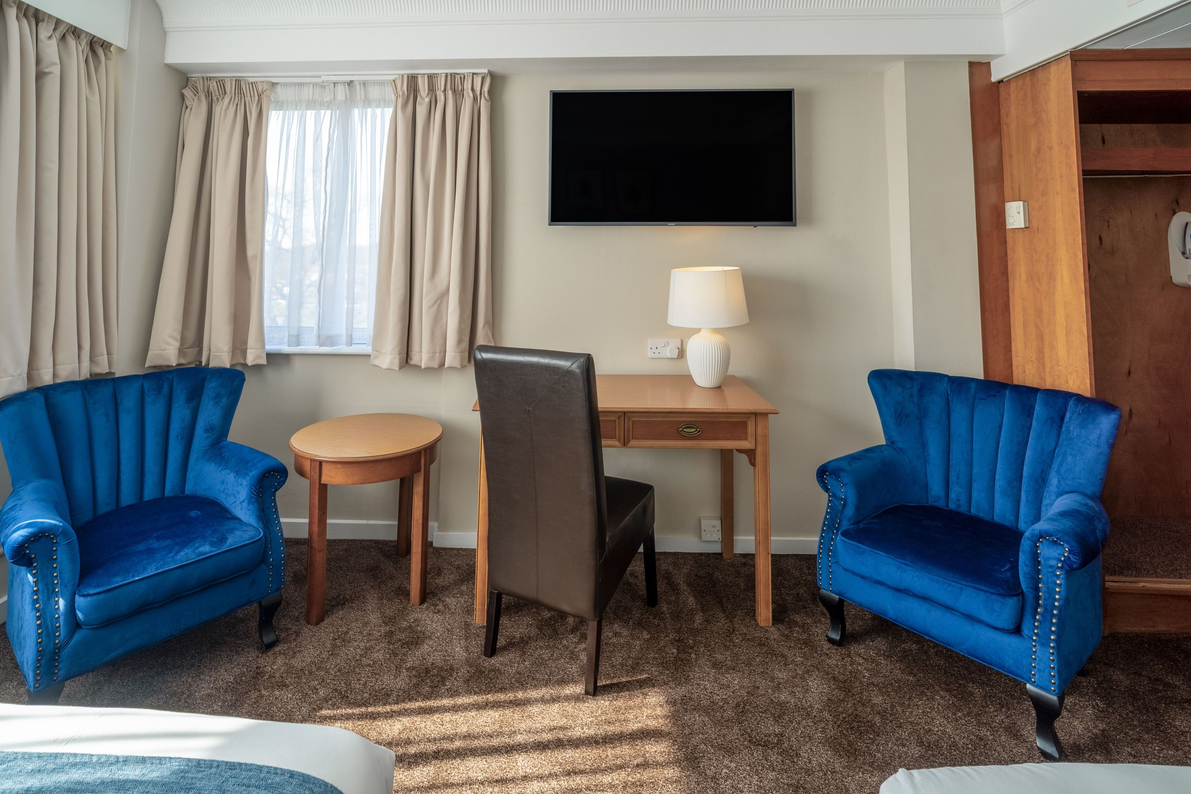 A hotel room with two blue armchairs, a small round table, a desk with a lamp, and a television on the wall.