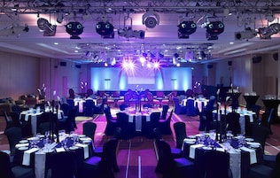 Uma sala de banquetes com mesas preparadas para um evento formal, equipada com iluminação de palco e sistemas de som.