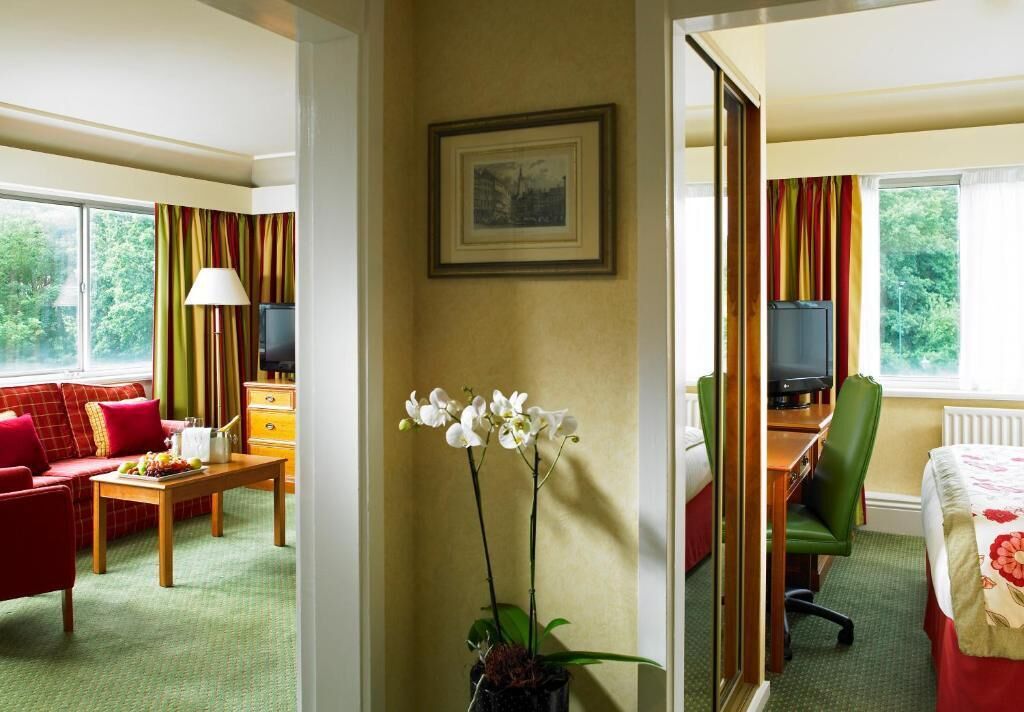 Quarto de hotel com sofá vermelho, escrivaninha de madeira, cadeira verde, televisão e janela com cortinas.