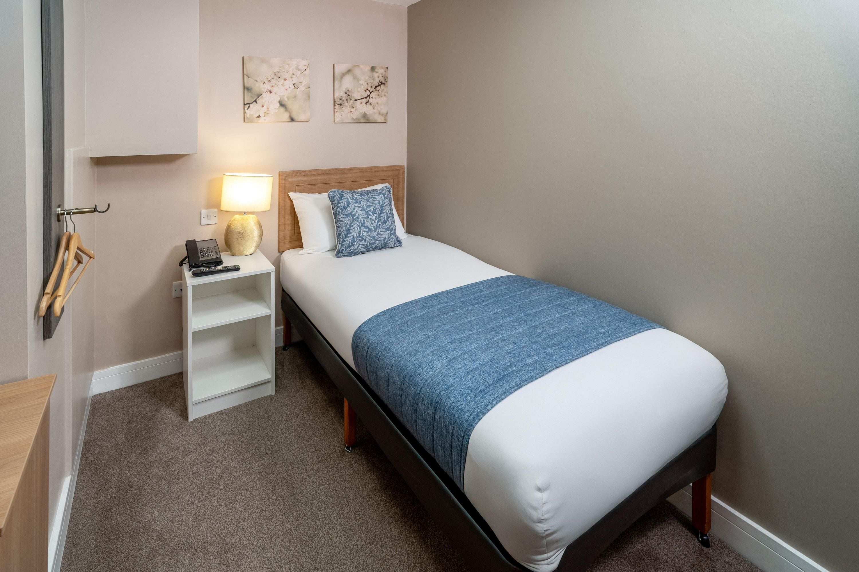 Une chambre d’hôtel avec un lit simple, une table de chevet avec une lampe, un téléphone et une étagère.