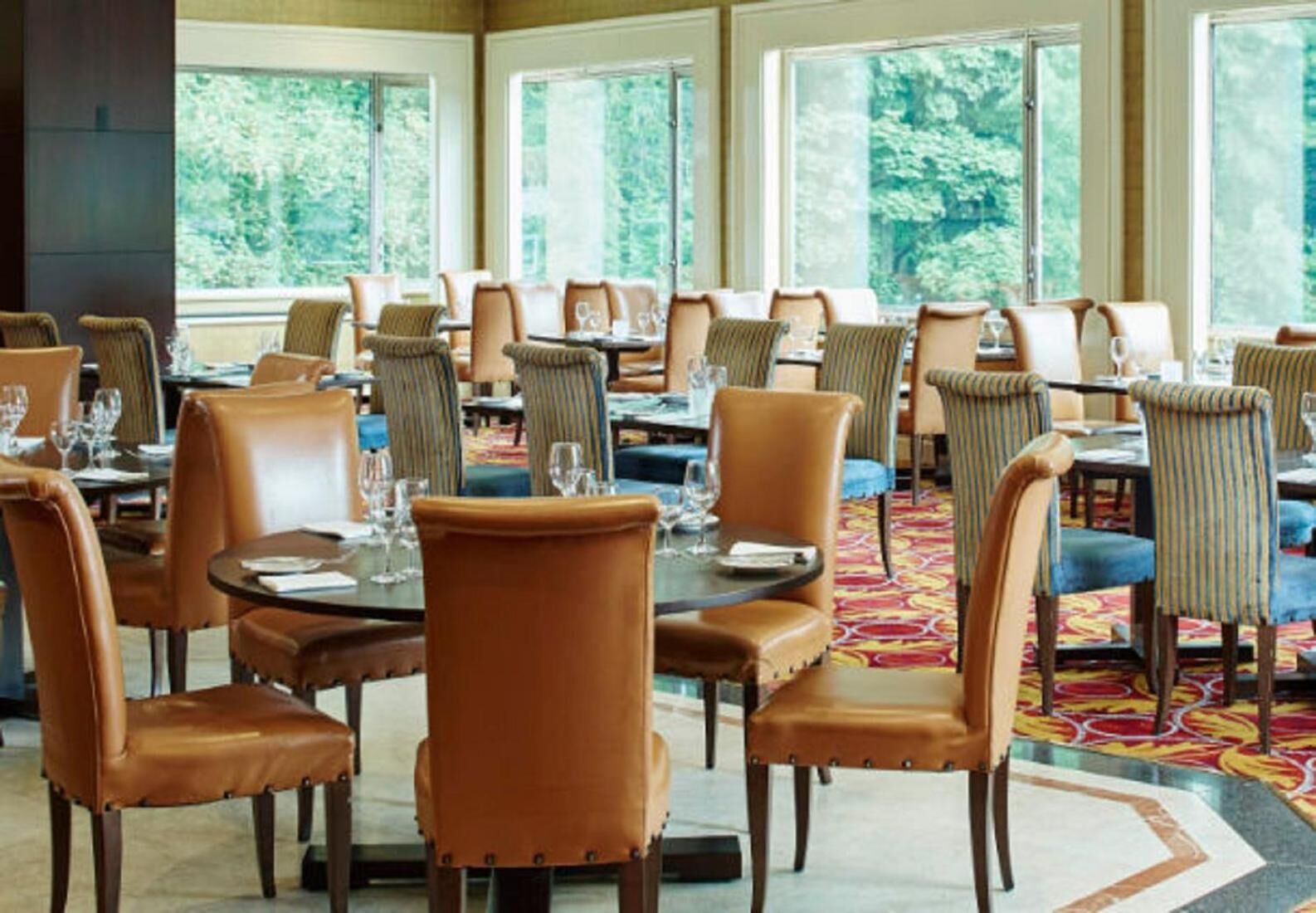 A dining area with round tables and chairs, large windows with a view of greenery, and a patterned carpet.