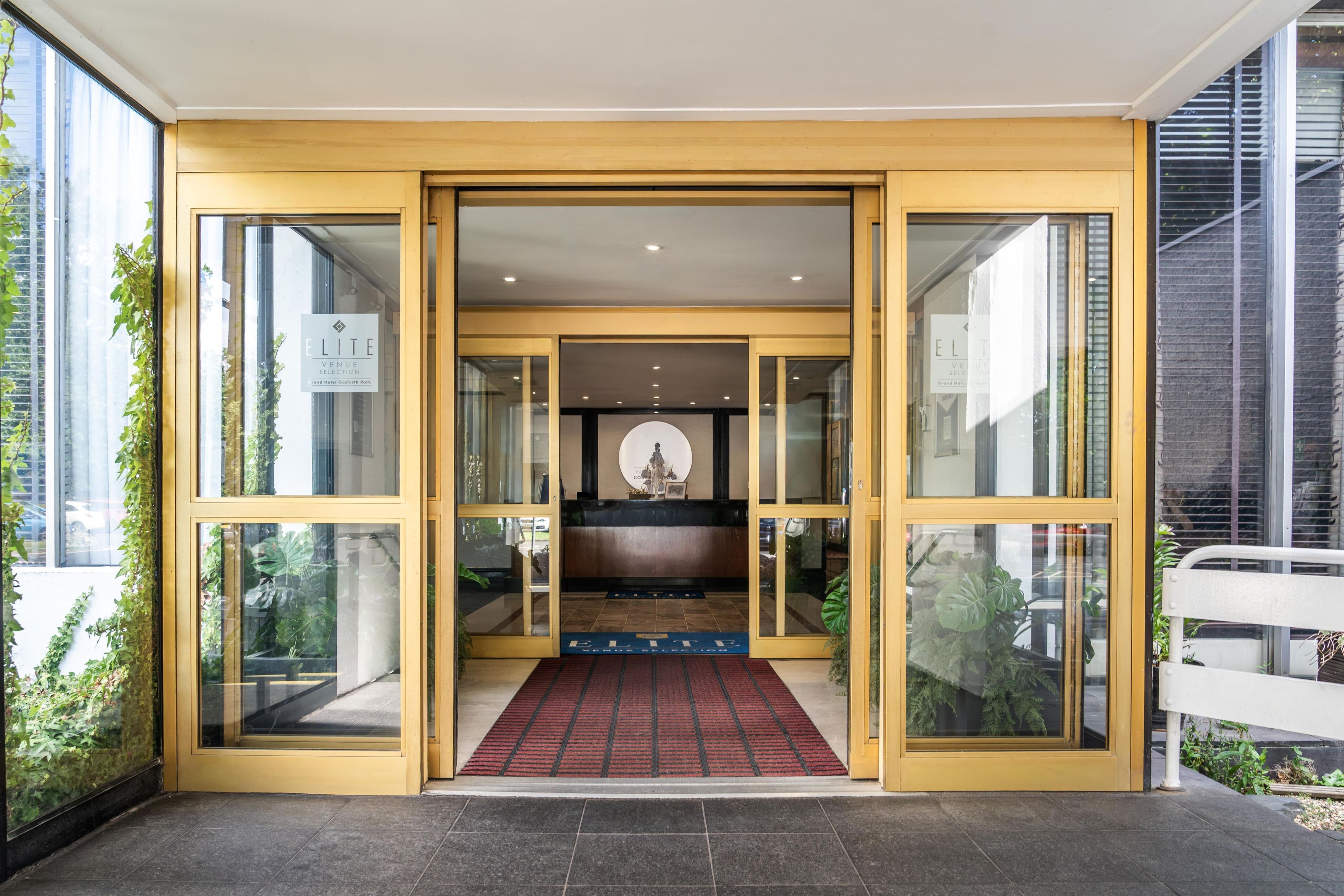 Entrada de hotel com portas de vidro, balcão de recepção e tapete vermelho.