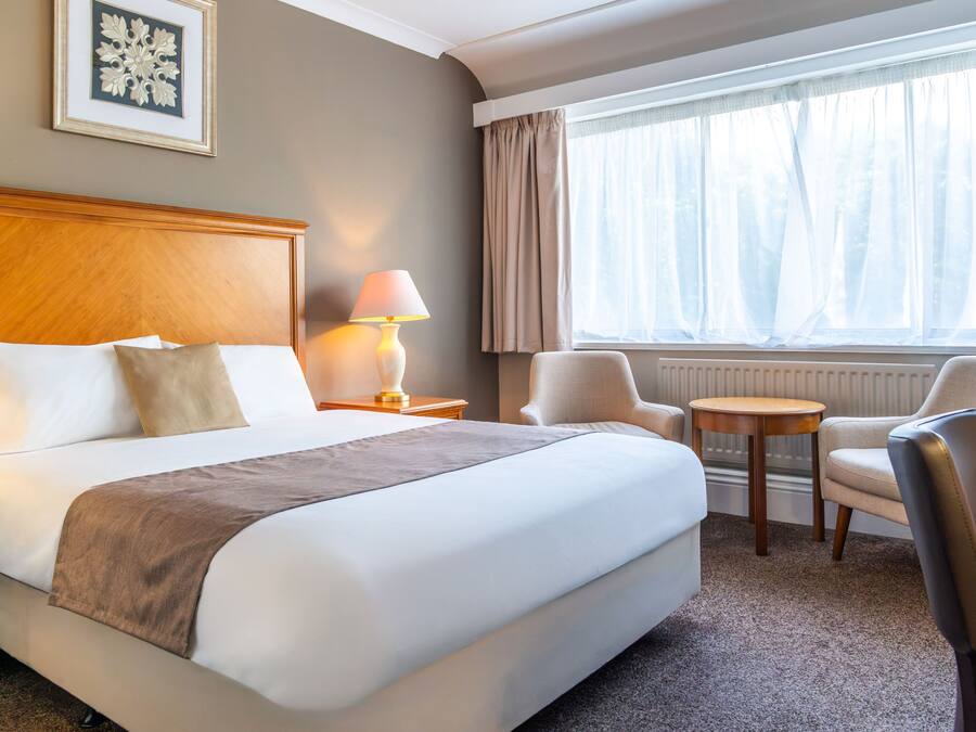 Une chambre d’hôtel avec un grand lit, une table de chevet, une lampe, une chaise et une fenêtre avec des rideaux.