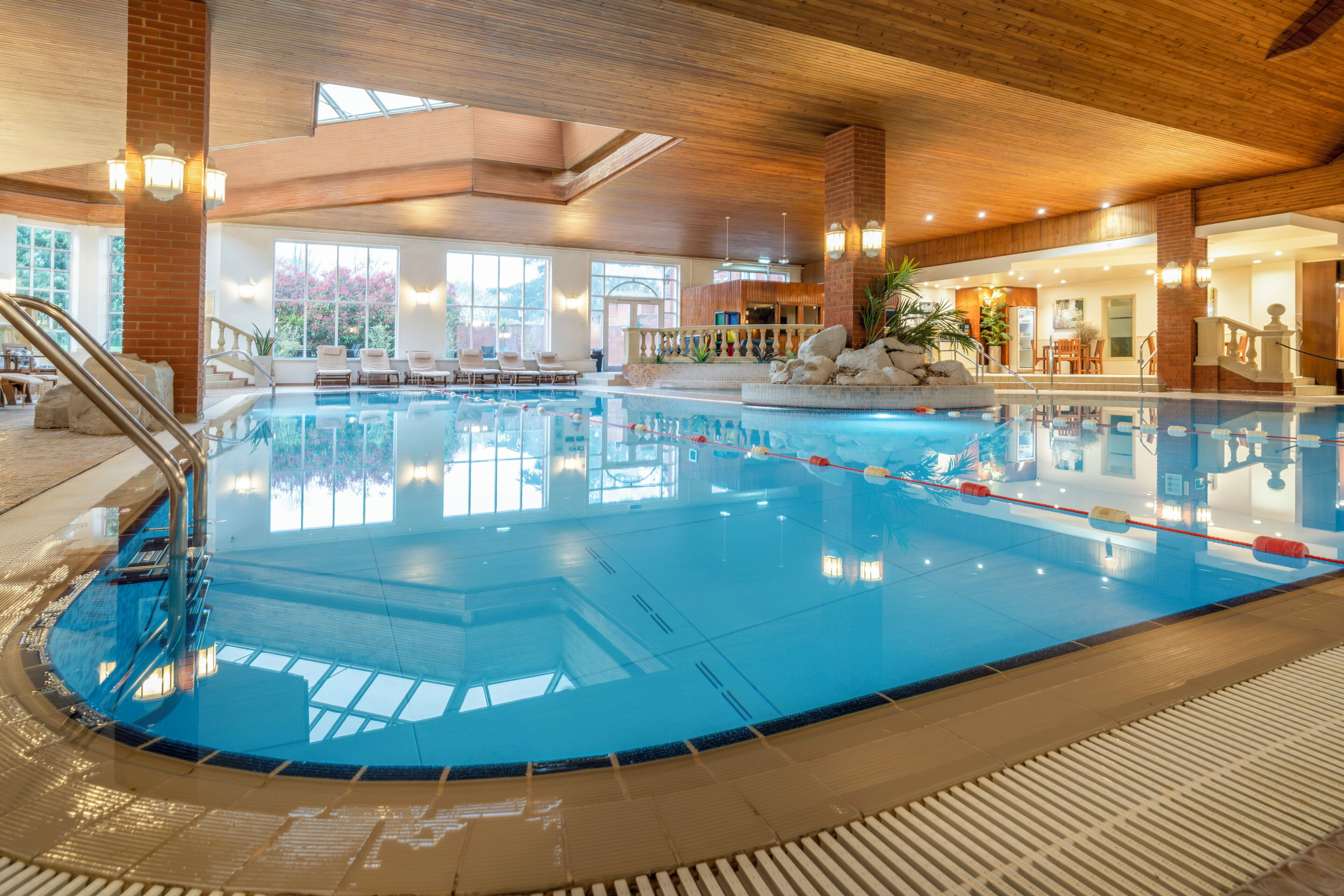 Piscina interior con techo de madera, ventanas de vidrio transparente y un área de estar con sillas y una mesa.