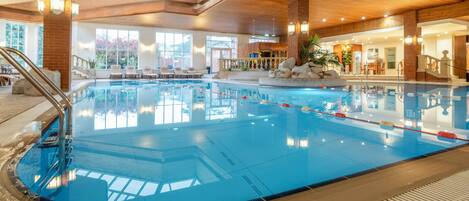 An indoor swimming pool with a wooden ceiling, clear glass windows, and a seating area with chairs and a table.