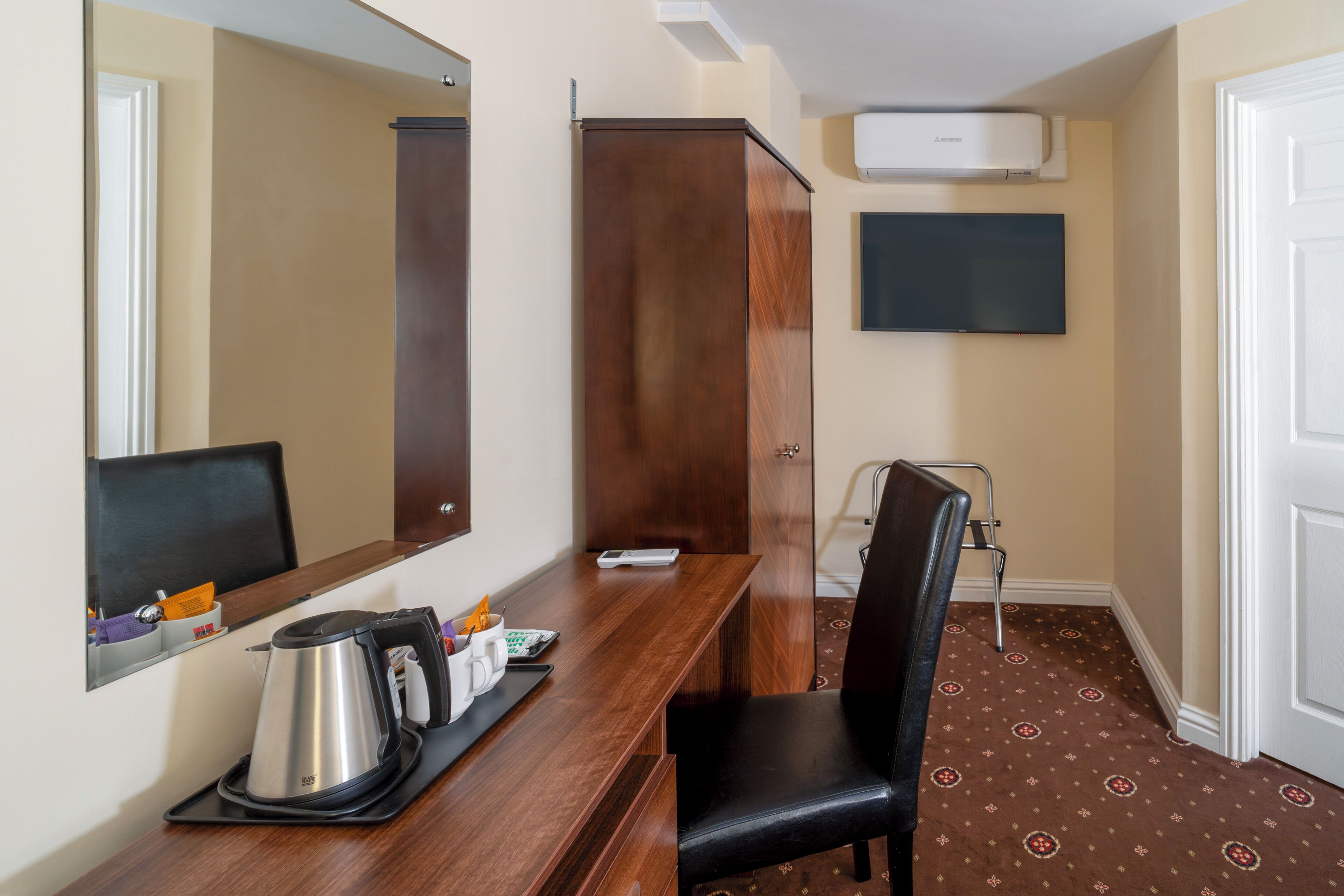 Ein Hotelzimmer mit Schreibtisch aus Holz, einem Sessel, einem großen Spiegel, einem flachbildfernseher an der Wand und einer Tür mit einem gemusterten Teppich.