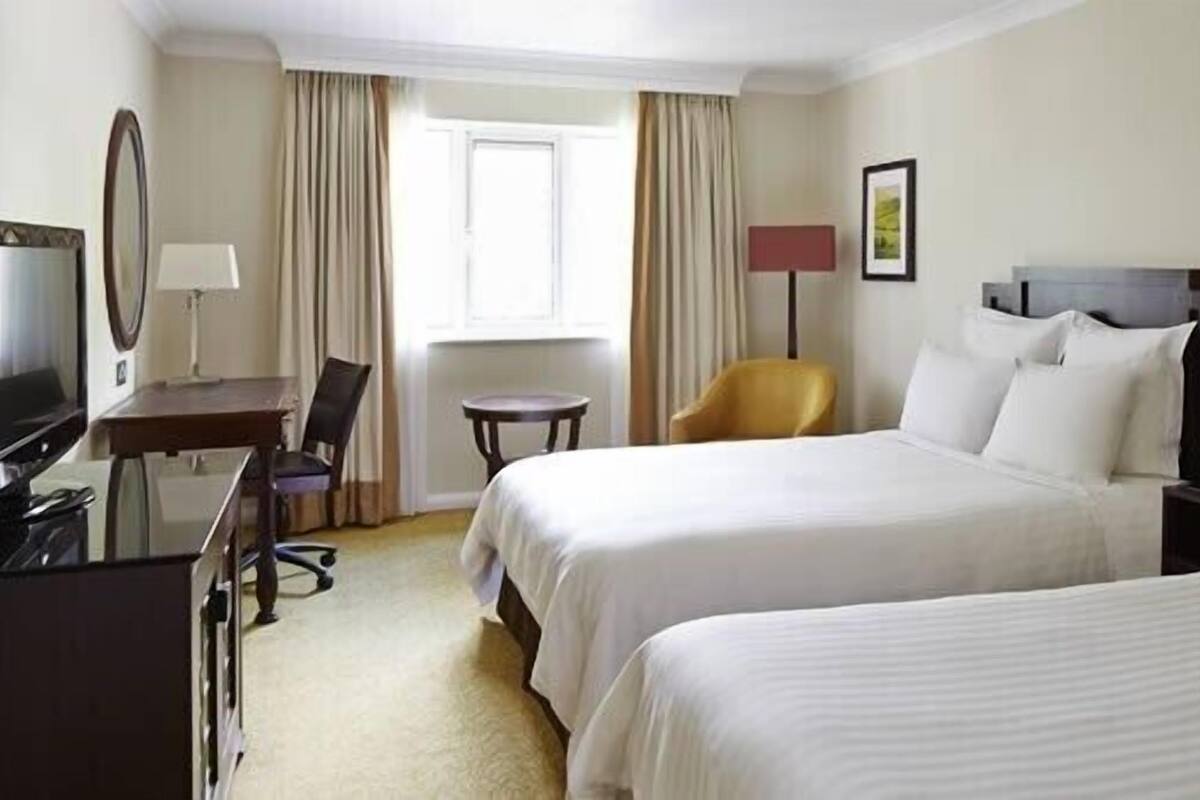 A hotel room with two beds, a desk with a computer, a chair, a lamp, and a window with curtains.