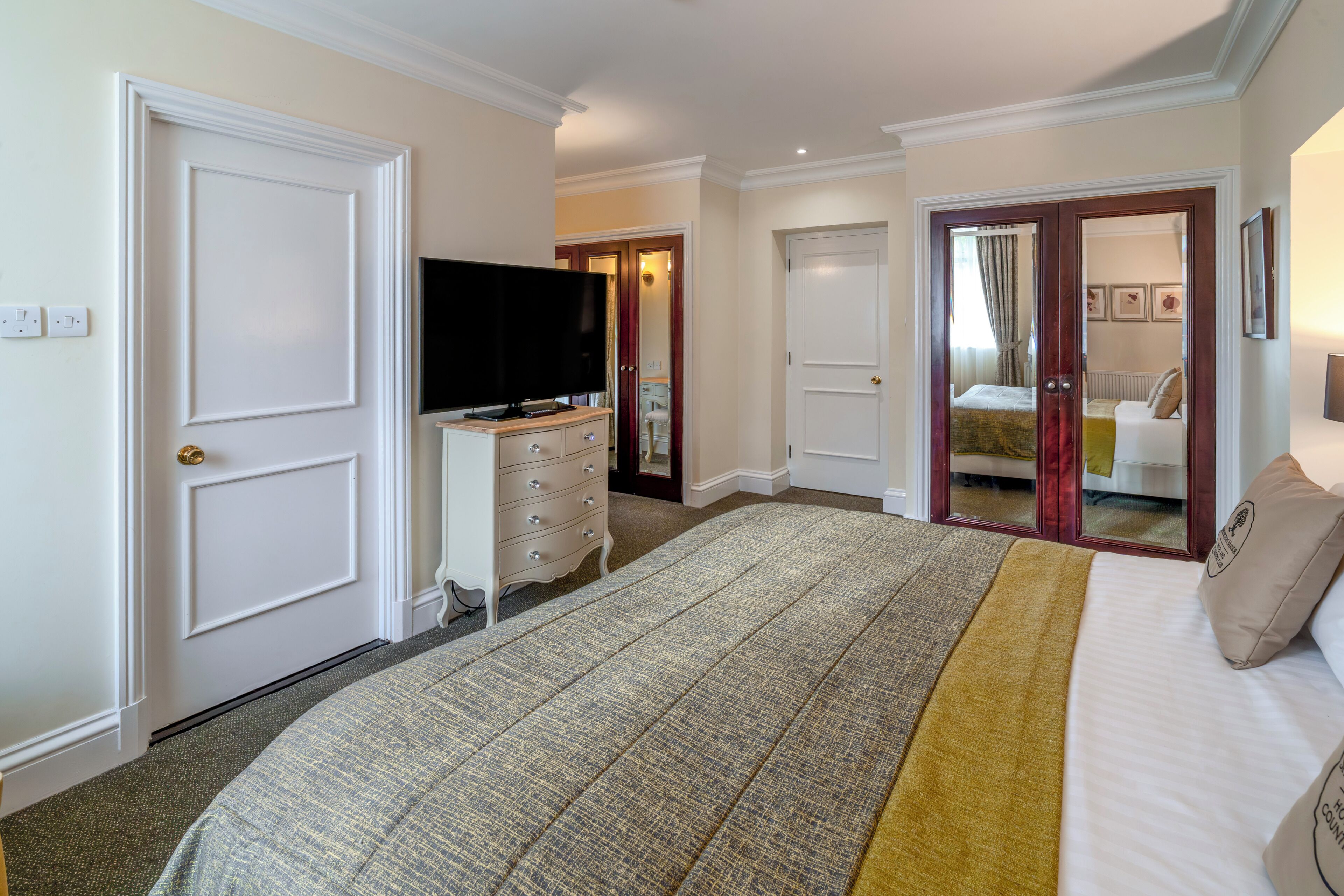 A hotel room with a large bed, a television, a dresser, and a door leading to another room.