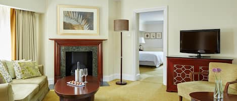 A hotel room with a sofa, a coffee table with a tray of snacks and glasses, a fireplace, a television, and a bed in the background.