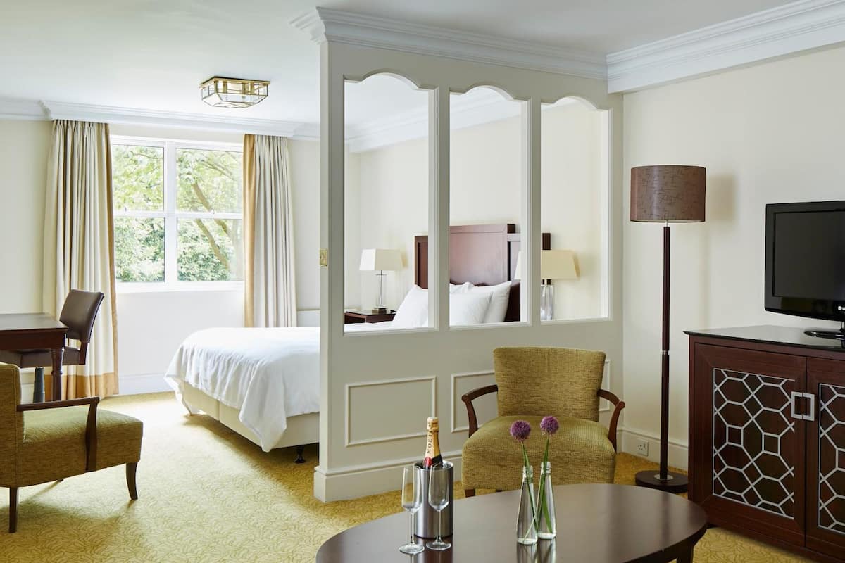 A hotel room with a four-poster bed, a sitting area with a table and chairs, a television, and a view of the outdoors through the window.
