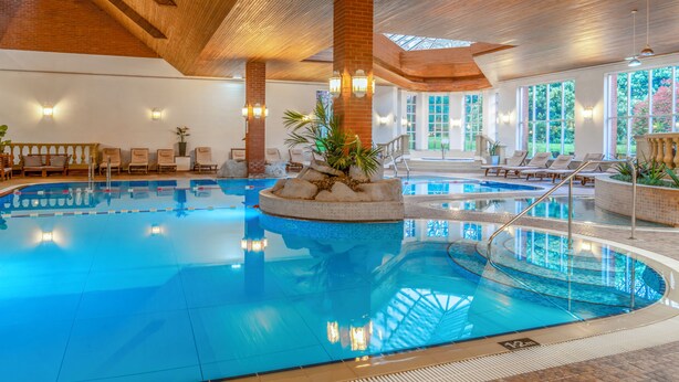 An indoor swimming pool with a central island, lounge chairs, and a wooden ceiling. - Sprowston Manor Hotel, Spa & Golf (Norwich)