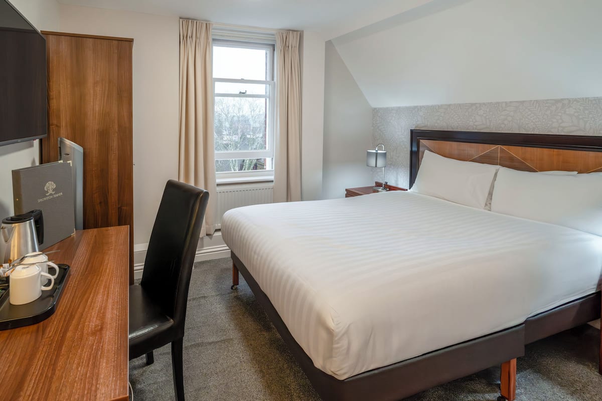 A hotel room with a bed, a desk with a coffee maker, a chair, a window with curtains, and a TV.