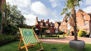 A green and white striped lounge chair in a well-maintained garden area with a brick building in the background. - Sprowston Manor Hotel, Spa & Golf (Norwich)