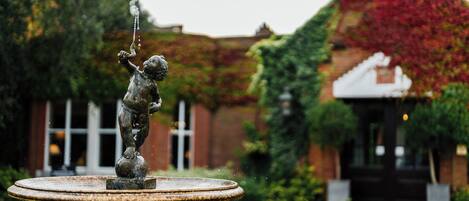 Uma fonte com uma estátua em um pátio, cercado por vegetação e um edifício com telhado vermelho.
