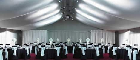 Une grande salle dotée d’un sol à carreaux, de rangées de tables recouvertes de nappes blanches et de chaises noires ornées de rubans blancs.