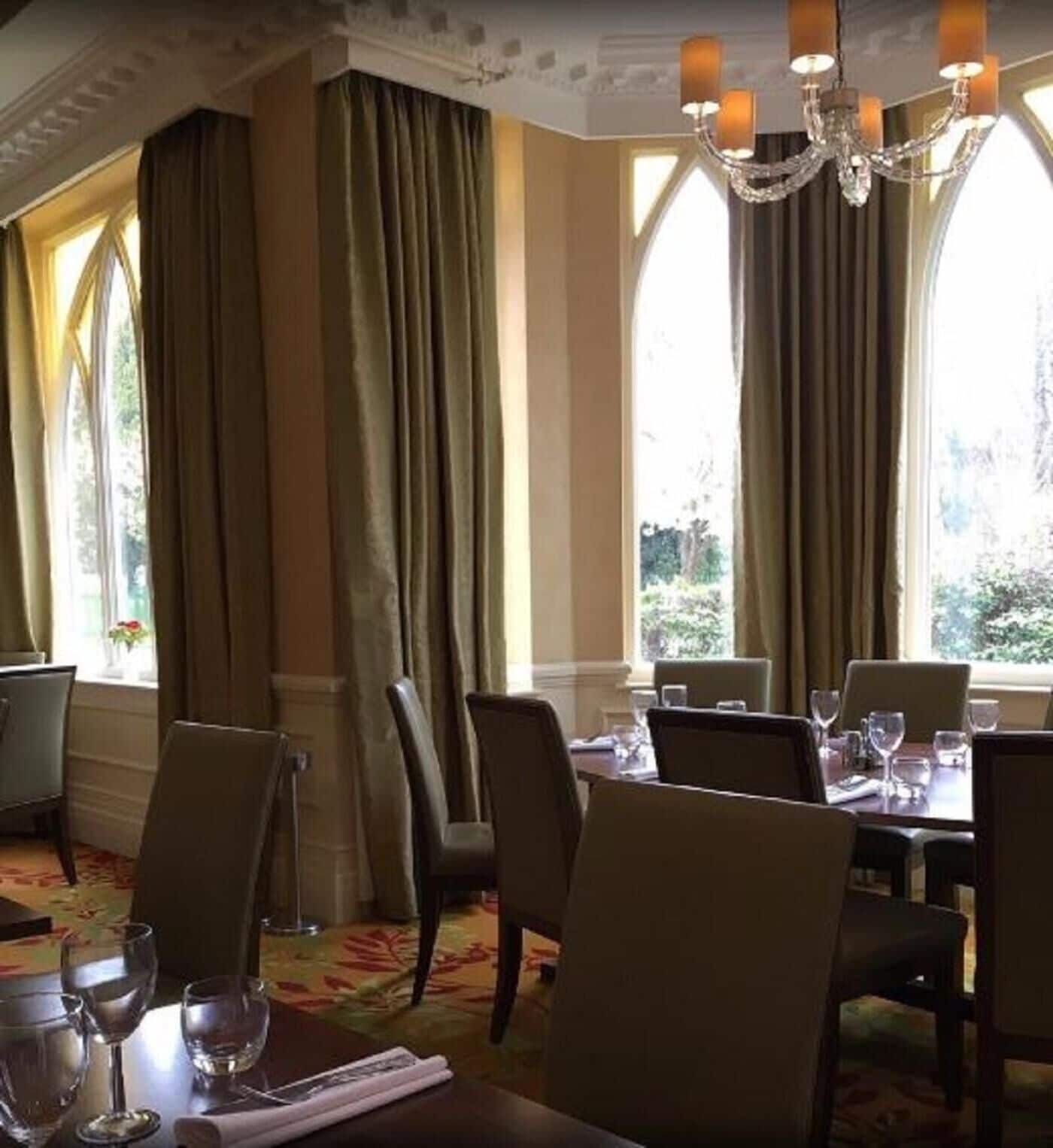 Comedor con mesas listas para el servicio, ventanas amplias con cortinas y una araña.