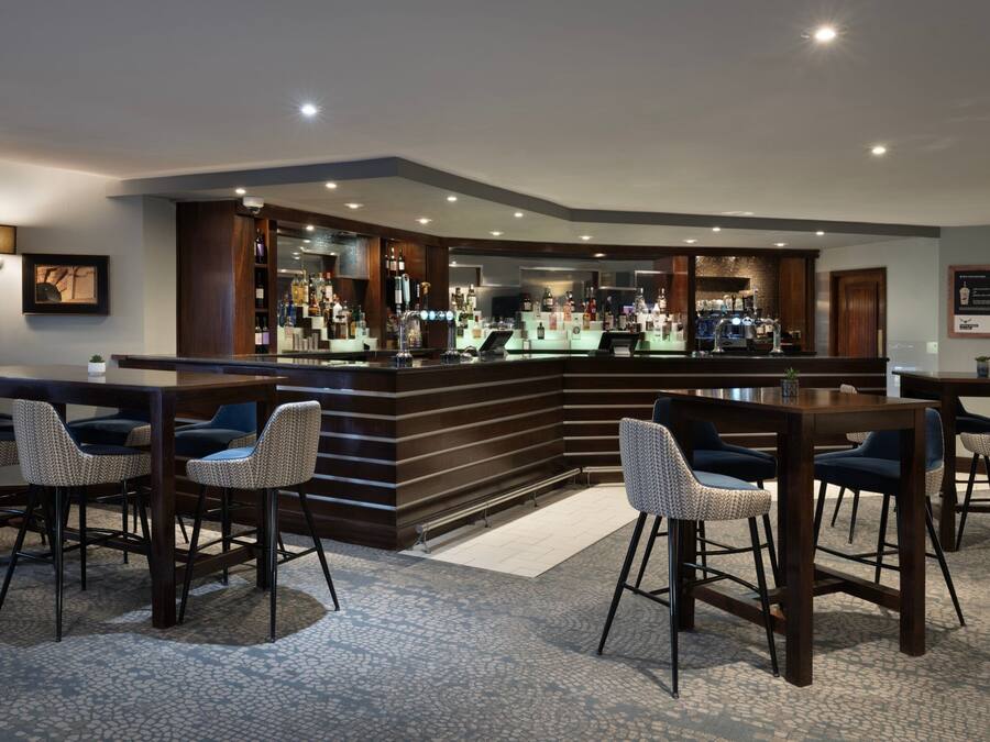 Zona de bar con una barra curva, taburetes y una variedad de botellas y vasos.