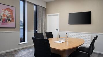 Un salón de conferencias con una mesa redonda, cuatro sillas negras, un televisor de pantalla plana, una ventana con cortinas y un radiador.