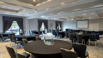 A conference room with round tables, chairs, a projector screen, and a large window with curtains.