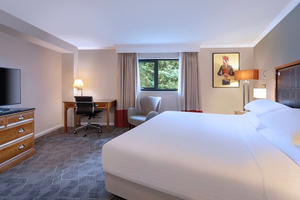 Habitación de hotel con una cama grande, un escritorio con silla, un televisor y un ventanal con cortinas.