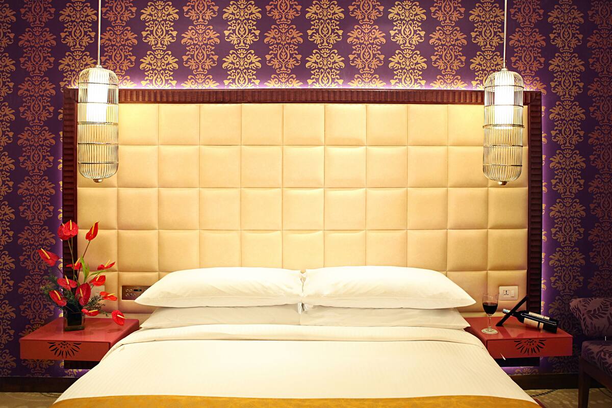 Une chambre d’hôtel avec une tête de lit capitonnée, un lit recouvert de draps blancs, une table de chevet ornée d’un vase de fleurs et deux lampes fixées au mur.
