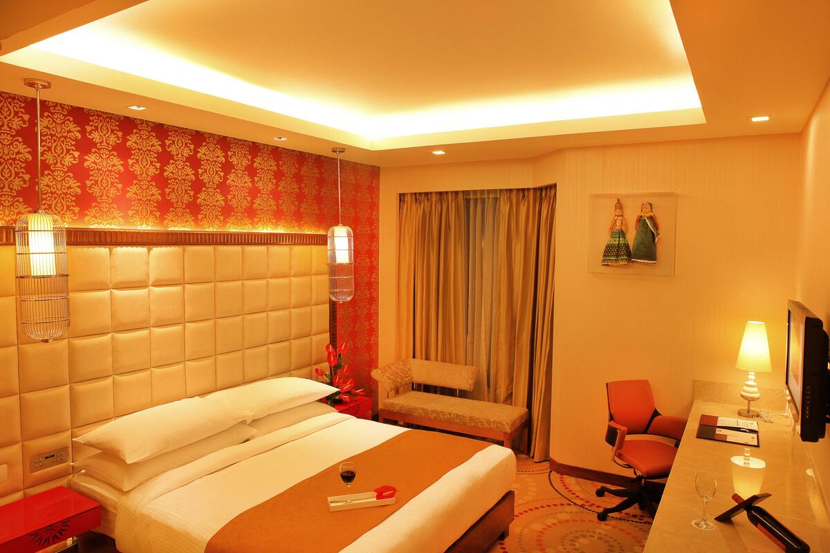Une chambre d’hôtel avec un grand lit, un bureau avec une lampe, une chaise, un téléviseur et une fenêtre avec des rideaux.
