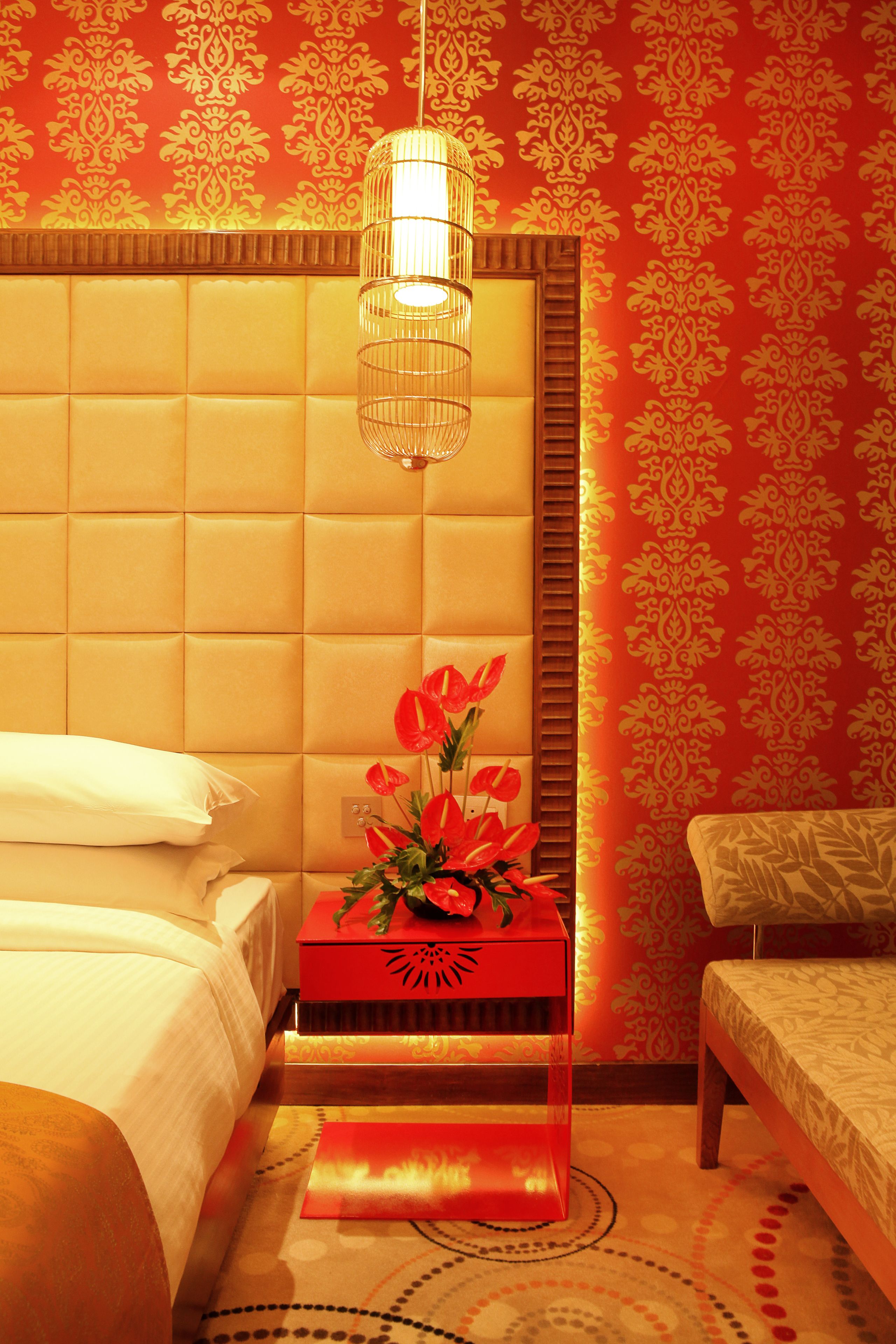 Una habitación de hotel con cabecera acolchada, mesita de noche con una caja roja y flores, y alfombra estampada.