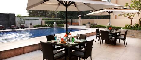 A poolside dining area with a table set for two, surrounded by chairs, and an umbrella providing shade.