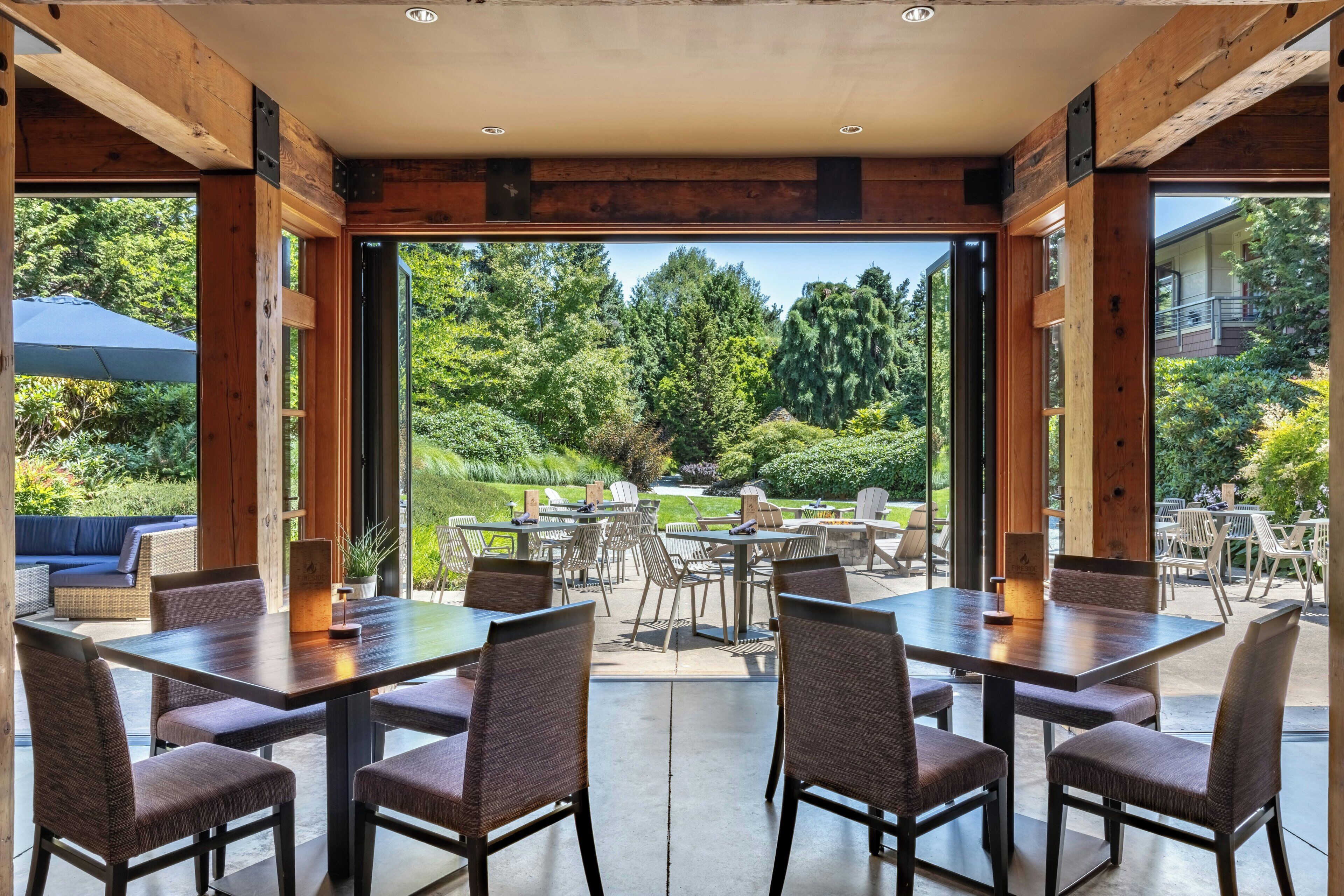A spacious outdoor dining area with multiple tables and chairs, large windows offering a view of a lush garden and trees.