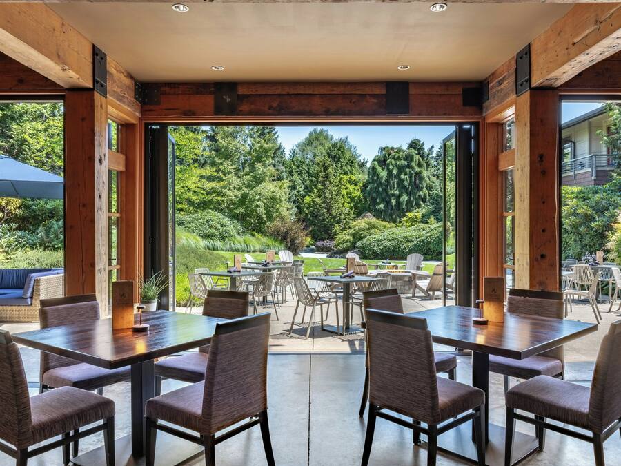 A spacious outdoor dining area with multiple tables and chairs, large windows offering a view of a lush garden and trees.