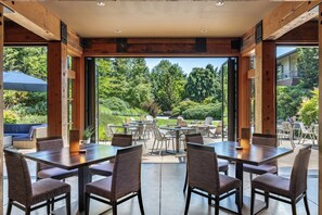 A spacious outdoor dining area with multiple tables and chairs, large windows offering a view of a lush garden and trees. - Willows Lodge (Woodinville)