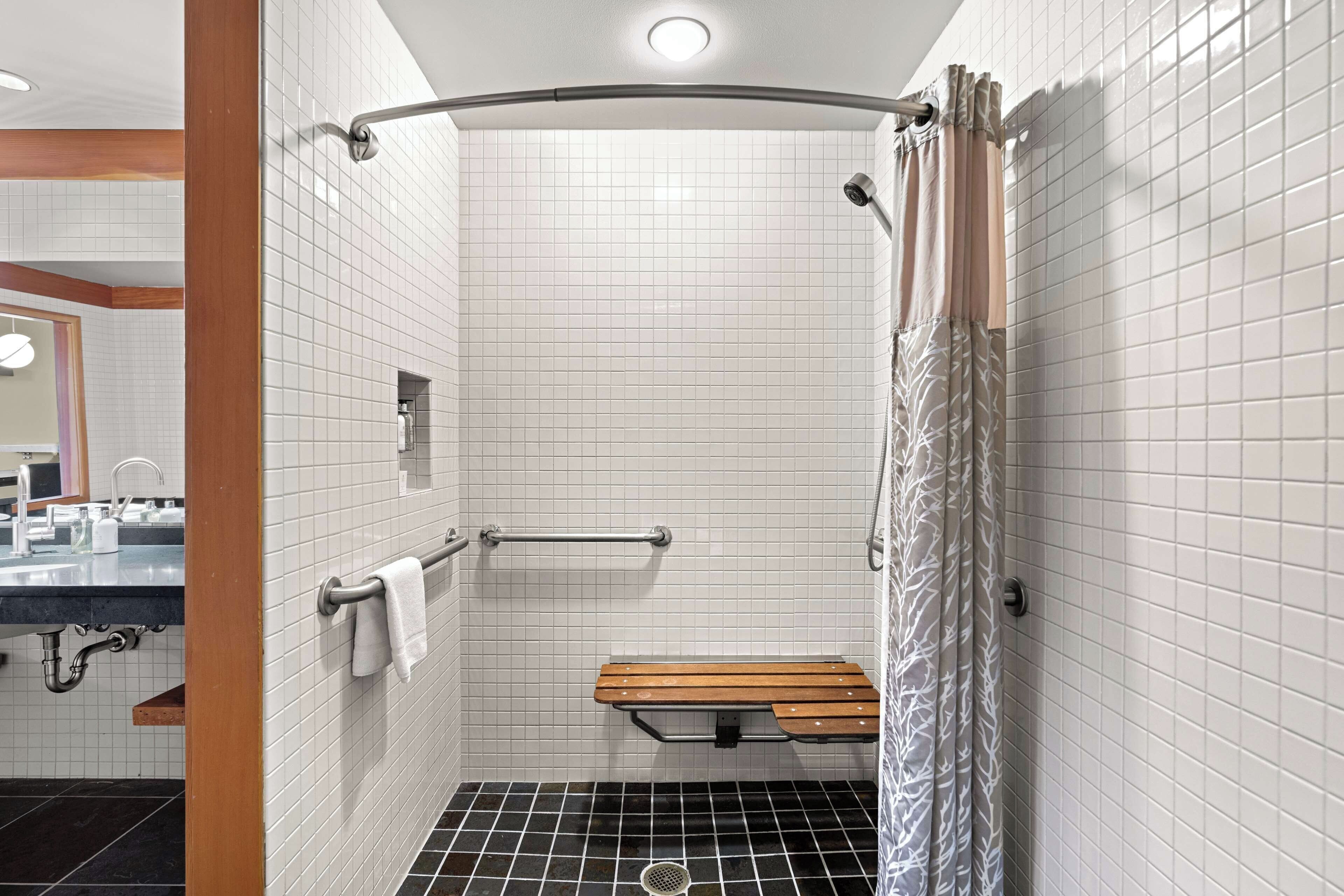 A modern bathroom with white tiled walls, a shower with a curved glass door, a wooden bench, and a handrail.