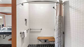 A modern bathroom with white tiled walls, a shower with a curved glass door, a wooden bench, and a handrail.