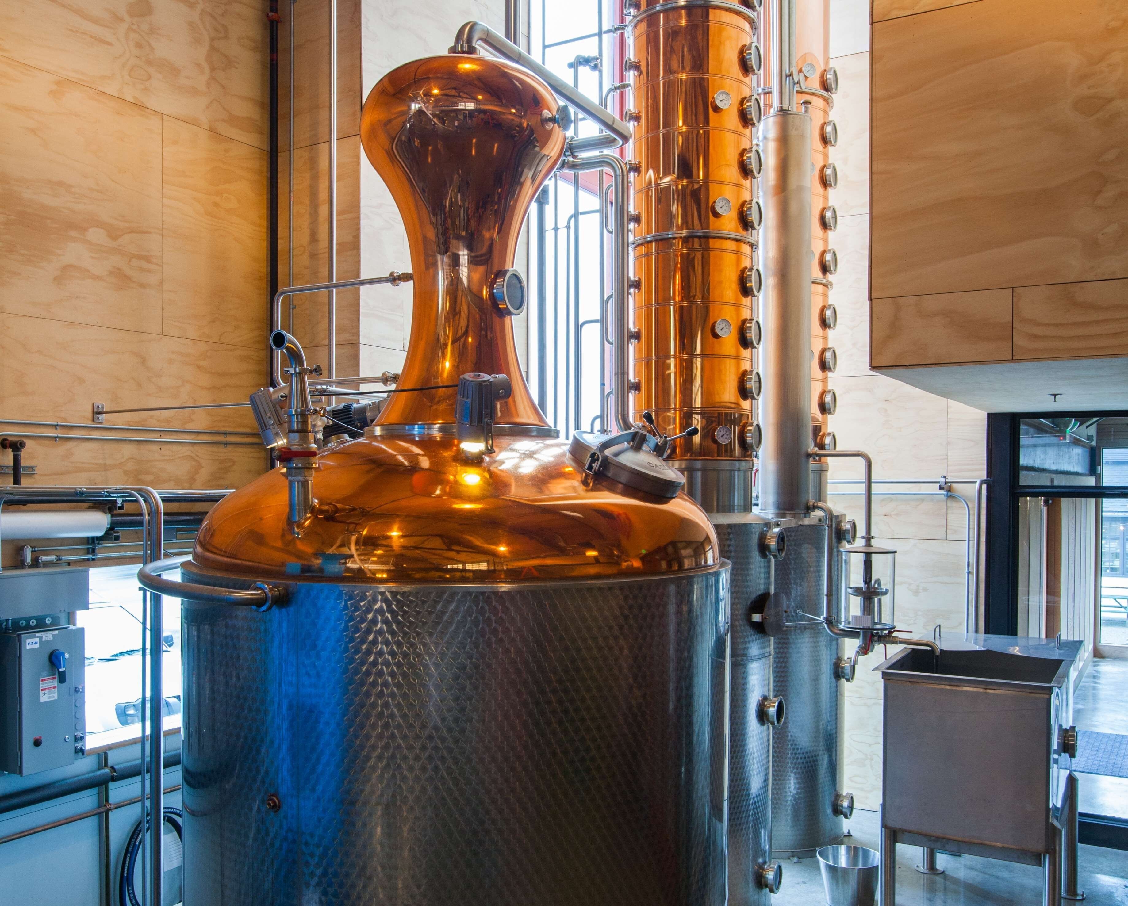A distillation apparatus with a copper still and a vertical column, surrounded by stainless steel tanks and pipes, in a modern industrial setting.