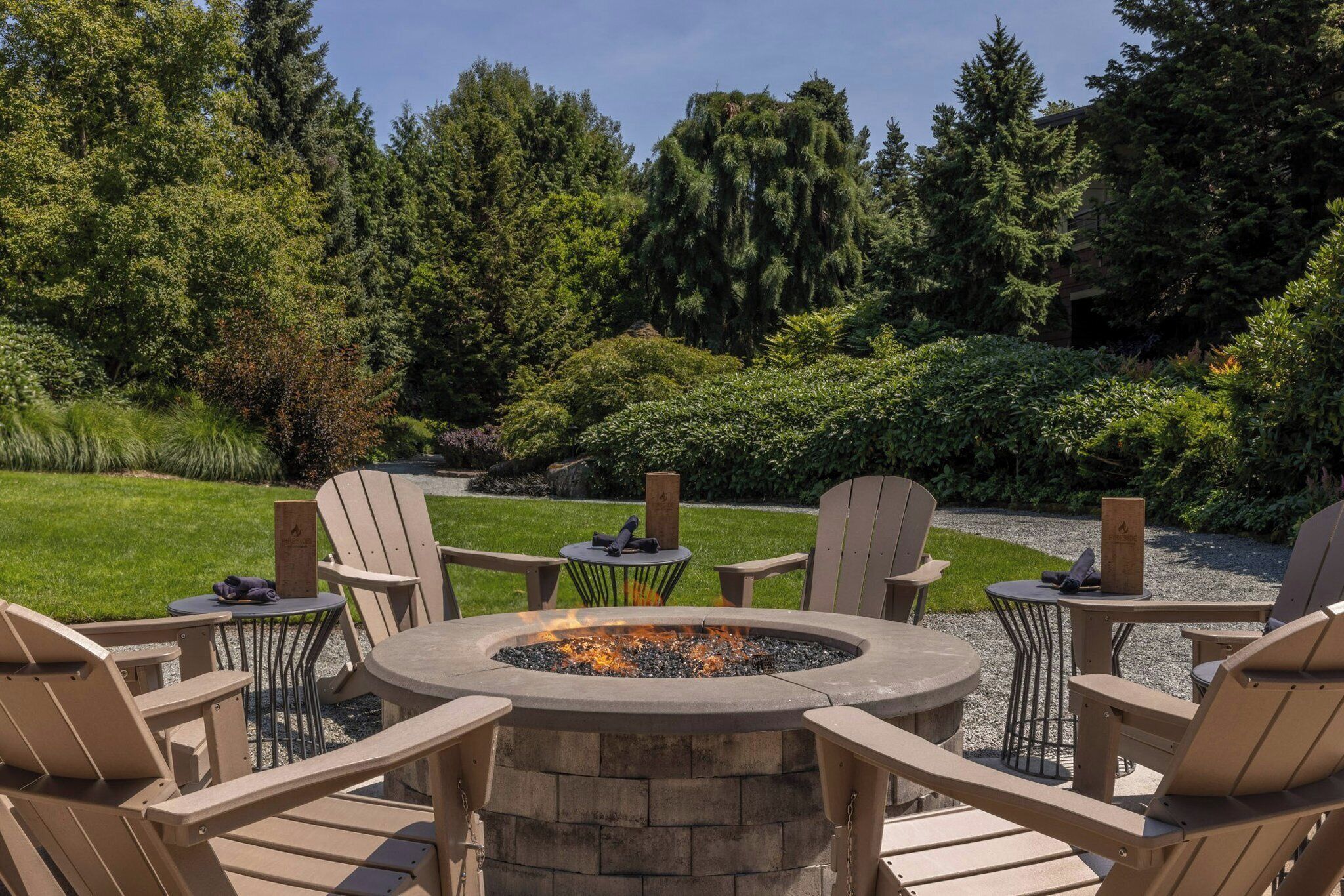 A fire pit surrounded by Adirondack chairs in a landscaped garden with trees and shrubs.
