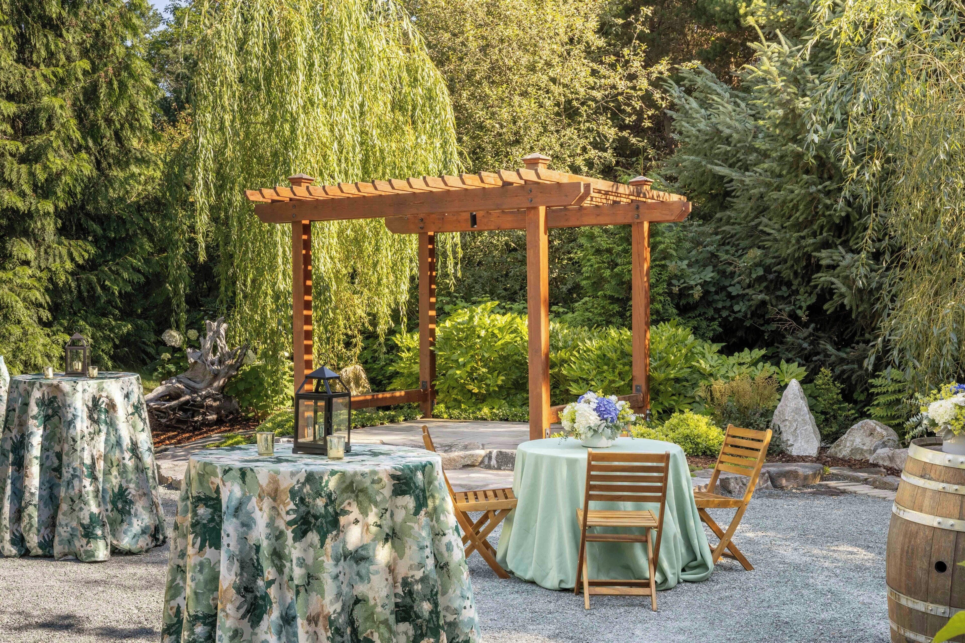 Un pabellón con techo de madera, rodeado de vegetación exuberante y árboles. Hay mesas con sillas dispuestas para comidas al aire libre, y un farol decorativo sobre una de las mesas.