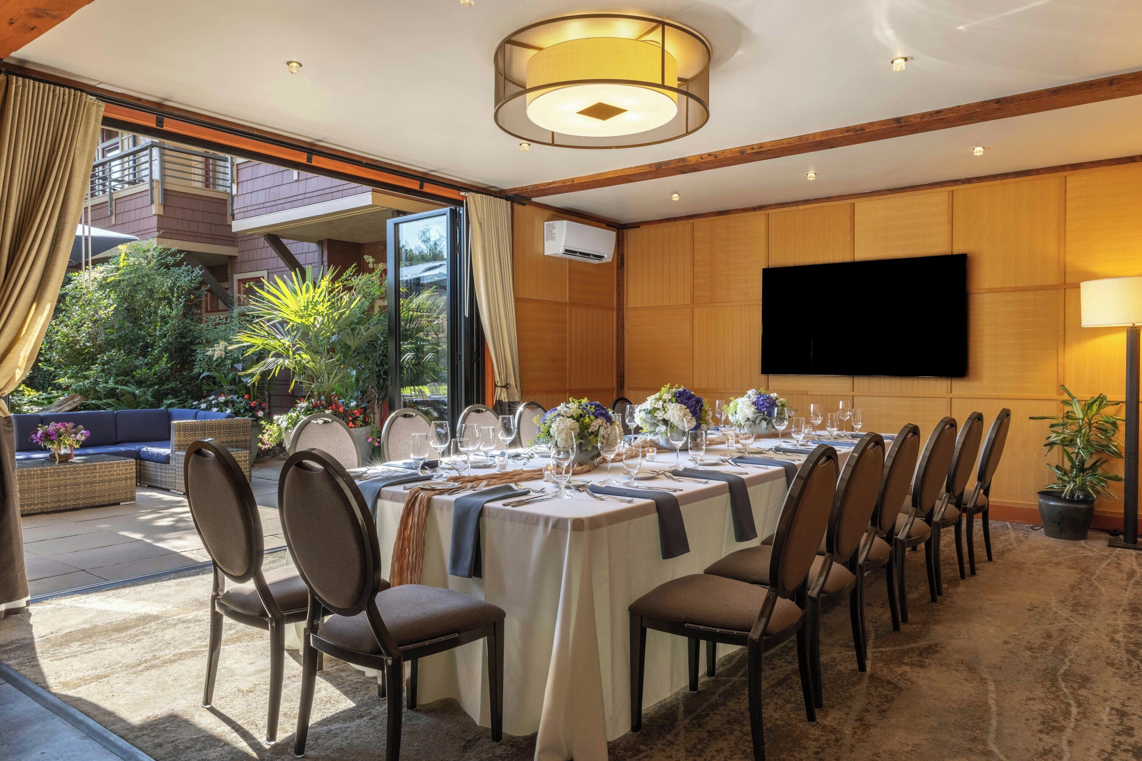 A dining area with a long table set for a meal, surrounded by chairs, with a television mounted on the wall and an outdoor view.