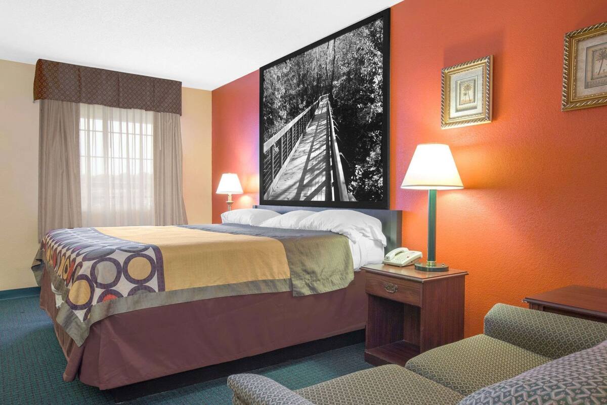 Habitación de hotel con cama, dos lámparas de noche, un teléfono, una silla y un cuadro grande enmarcado en la pared.