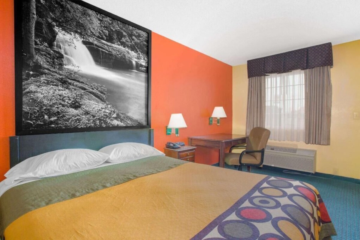 A hotel room with a bed, two bedside lamps, a desk, a chair, a large framed photo, and a window with curtains.