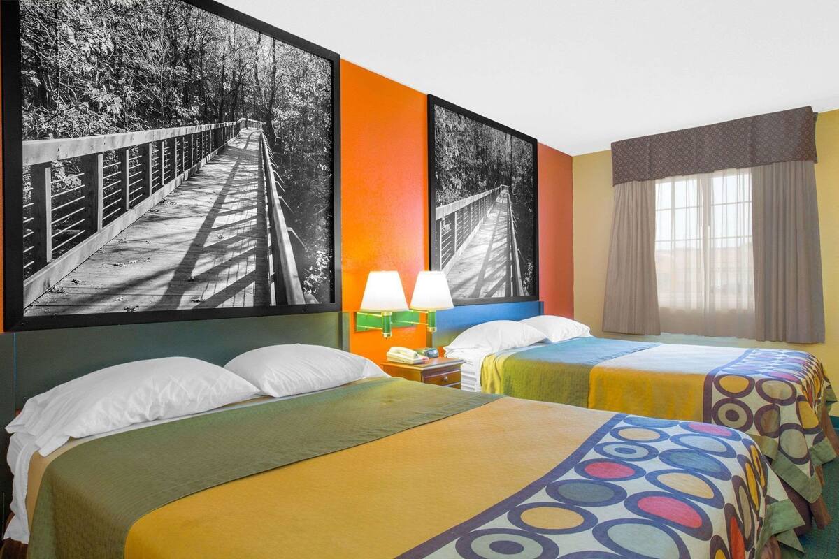 Habitación de hotel con dos camas, una mesita de noche con lámpara y cuadros grandes enmarcados en las paredes.