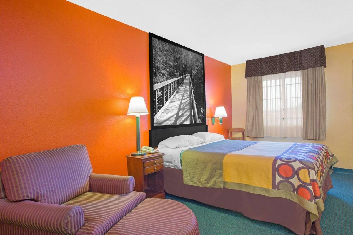 Habitación de hotel con una cama, una silla, una mesita de noche, una lámpara, una ventana con cortinas y una gran obra de arte en la pared.
