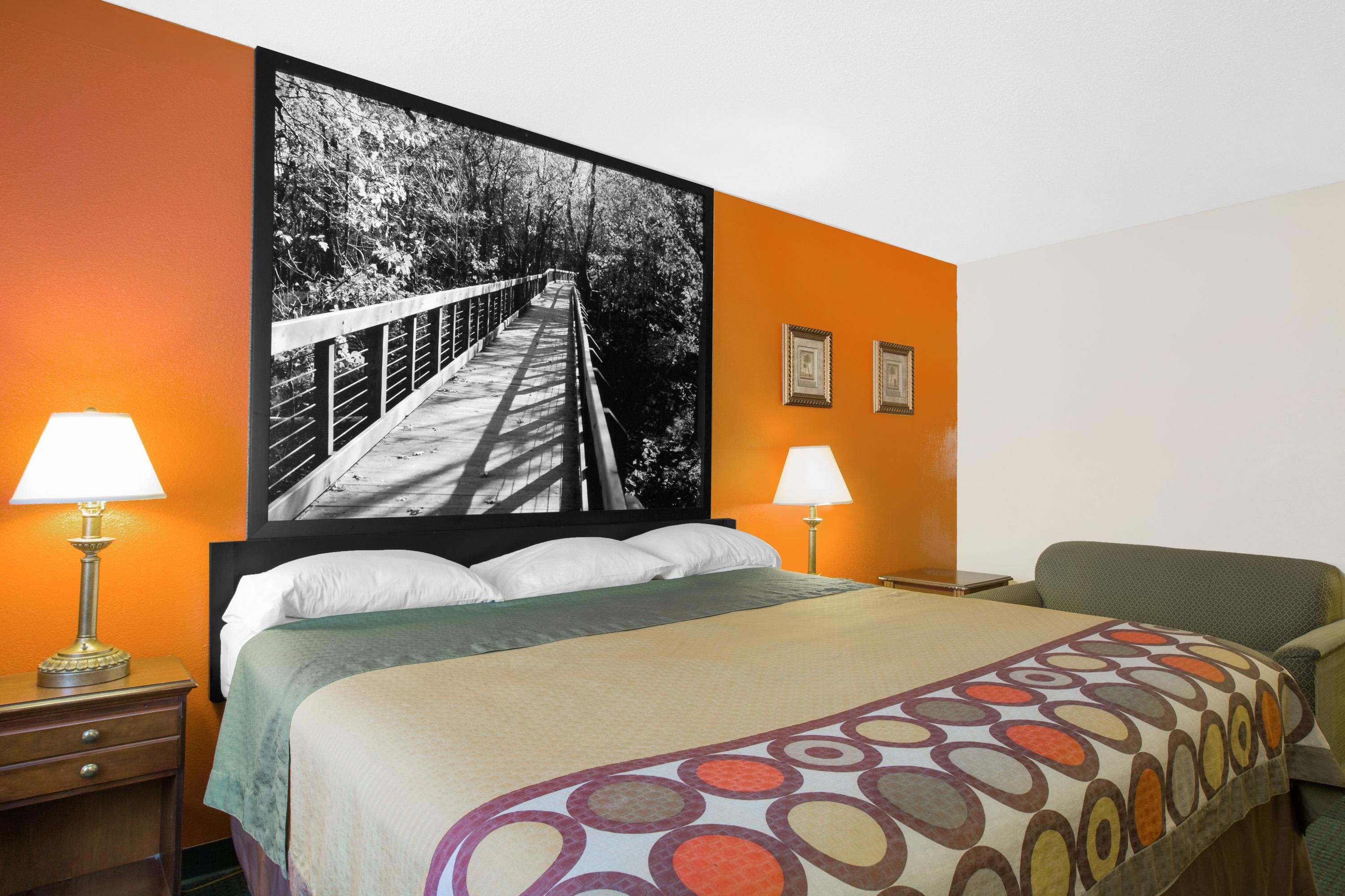 A hotel room with a bed, two bedside lamps, a chair, and a large framed photograph on the wall.