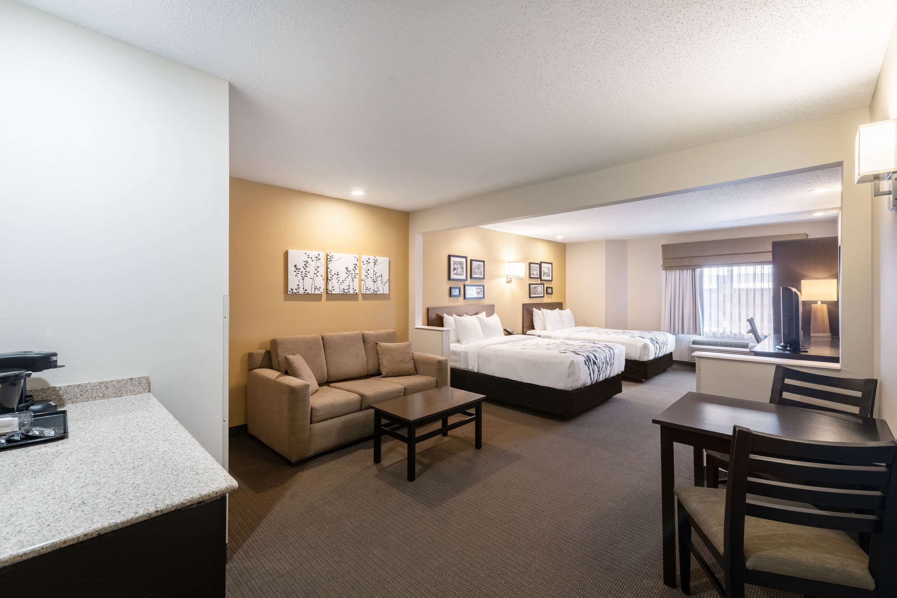 Ein Hotelzimmer mit einer Couch, einem kleinen Tisch, einem Bett und einem Schreibtisch. An den Wänden hängen Bilder in Rahmen und es gibt ein Fenster mit Vorhängen.