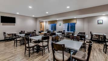 A hotel dining room with multiple tables and chairs, a flat-screen TV mounted on the wall, and windows with curtains.