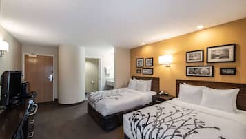 A hotel room with two beds, a desk with a computer, and a TV. There are framed pictures on the wall and a lamp on the desk.